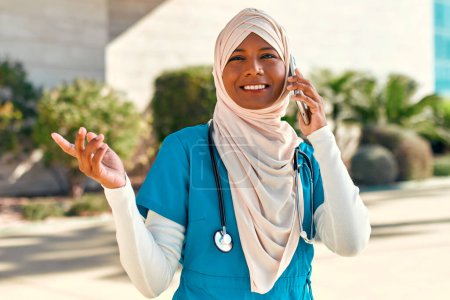 Foto de Mujer musulmana joven en el médico de hijab en uniforme con un estetoscopio hablando por teléfono mientras está de pie en la calle cerca del edificio del hospital. Medicina y asistencia sanitaria. - Imagen libre de derechos
