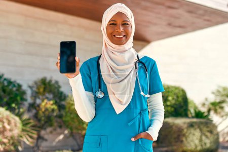 Foto de Mujer musulmana joven en hijab médico en uniforme con estetoscopio que muestra la pantalla del teléfono inteligente mientras está de pie en la calle cerca del edificio del hospital. Medicina y asistencia sanitaria. - Imagen libre de derechos