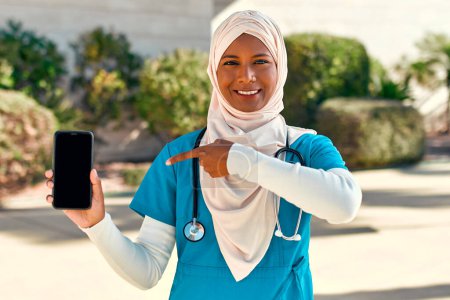 Téléchargez les photos : Jeune femme musulmane en hijab médecin en uniforme avec stéthoscope montrant écran smartphone tout en se tenant dans la rue près du bâtiment de l'hôpital. Médecine et soins de santé. - en image libre de droit