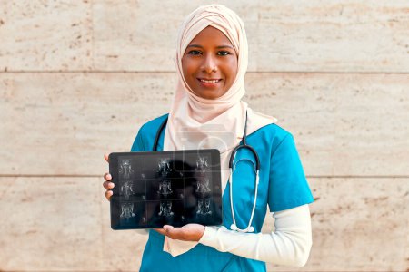 Foto de Mujer musulmana joven en el médico de hijab en uniforme con un estetoscopio sosteniendo una tableta con rayos X de pie en la calle cerca del edificio del hospital. Medicina y asistencia sanitaria. - Imagen libre de derechos