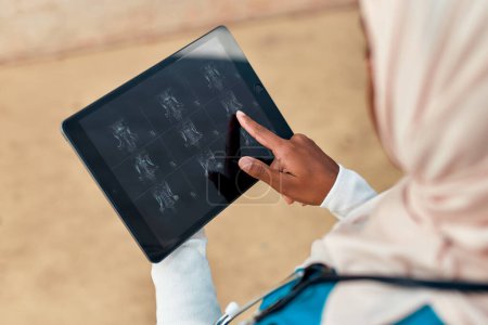 Foto de Mujer musulmana joven en el médico de hijab en uniforme con un estetoscopio sosteniendo una tableta con rayos X de pie en la calle cerca del edificio del hospital. Medicina y asistencia sanitaria. - Imagen libre de derechos