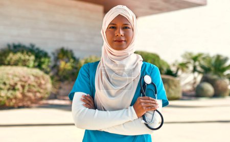 Photo for A young Muslim woman, a doctor in a hijab in uniform with a stethoscope, is standing on the street outside the hospital. Medicine and health care. - Royalty Free Image