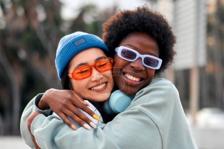 Téléchargez les photos : Afro-américain et asiatique jeunes femmes mignonnes copines câlins et avoir du plaisir à l'extérieur en écoutant de la musique dans les écouteurs. - en image libre de droit