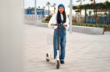 Photo for Pretty Asian young woman in a hat riding a scooter on the streets of the city. Active recreation and sports. - Royalty Free Image