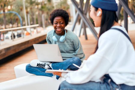 Foto de Mujeres jóvenes afroamericanas y asiáticas sentadas con piernas cruzadas al aire libre con computadora portátil y tableta. Freelancing y ganar en línea. - Imagen libre de derechos
