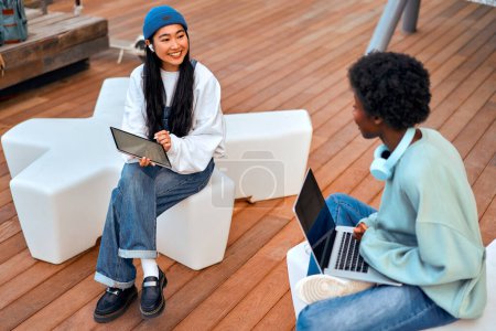 Photo for Young African American and Asian women sitting cross-legged outdoors with laptop and tablet. Freelancing and earning online. - Royalty Free Image