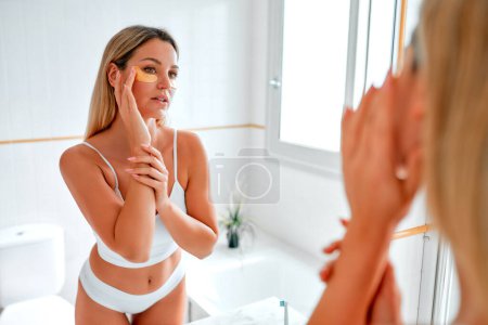 Téléchargez les photos : Une femme blonde en lingerie blanche debout dans la salle de bain au miroir, faisant sa routine matinale pour les soins de la peau du visage et du corps en utilisant des patchs pour les yeux. Procédures de cosmétologie et spa. - en image libre de droit