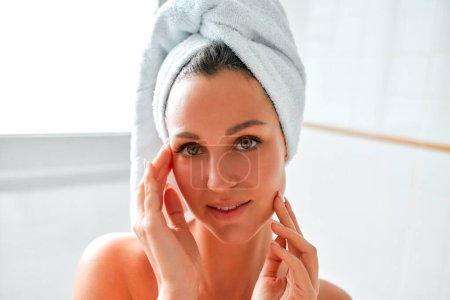 Téléchargez les photos : Une femme dans une serviette debout dans la salle de bain faisant sa routine matinale pour les soins de la peau du visage et du corps. Procédures de cosmétologie et spa. - en image libre de droit