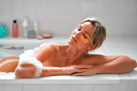 Photo for Beautiful woman lying in a white foam bath, bathing, enjoying and relaxing, using eye mask patches. Morning routine. Cosmetology and spa procedures. - Royalty Free Image