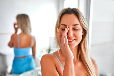Téléchargez les photos : Une femme blonde en lingerie bleue debout dans la salle de bain au miroir, faisant sa routine matinale pour les soins du visage et du corps. Procédures de cosmétologie et spa. - en image libre de droit