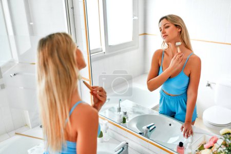 Foto de Hermosa mujer en lencería de pie en el lavabo y espejo haciendo masaje facial con un rodillo. Rutina de cuidado de la piel. Procedimientos de cosmetología y spa. - Imagen libre de derechos