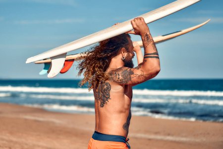 Photo for A couple of surfers on the beach. Caucasian woman in a swimsuit and a bearded buff man with tattoos in swimming trunks with surfboards near the sea. Sports and active recreation. - Royalty Free Image