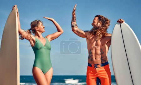 Téléchargez les photos : Femme caucasienne en maillot de bain et un homme musclé barbu avec des tatouages dans des maillots de bain avec des planches de surf près de la mer donnant un geste cinq haut. Sports et loisirs actifs. - en image libre de droit