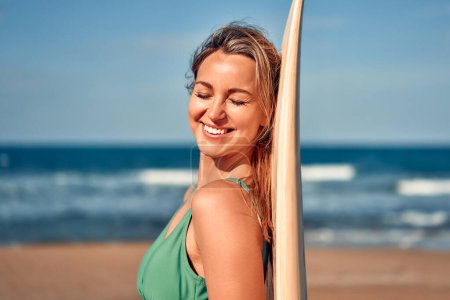 Téléchargez les photos : Belle femme caucasienne en maillot de bain avec une planche de surf debout sur une plage de sable près de la mer. Sports et loisirs actifs. - en image libre de droit