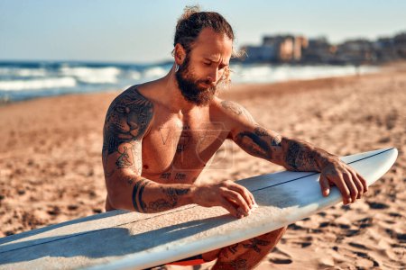 Foto de Caucásico hombre barbudo fornido con tatuajes en trajes de baño está encerando una tabla de surf cerca del mar. Deportes y recreación activa. - Imagen libre de derechos