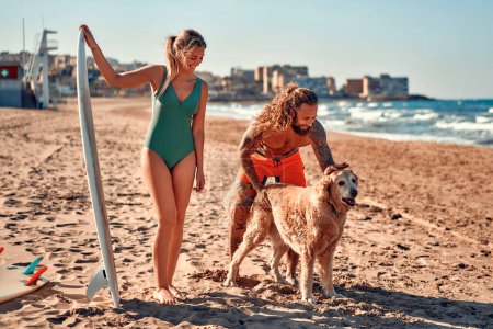 Photo for A couple of surfers with a dog on the beach. Caucasian woman in a swimsuit and a bearded buff man with tattoos in swimming trunks with surfboards near the sea. Sports and active recreation. - Royalty Free Image