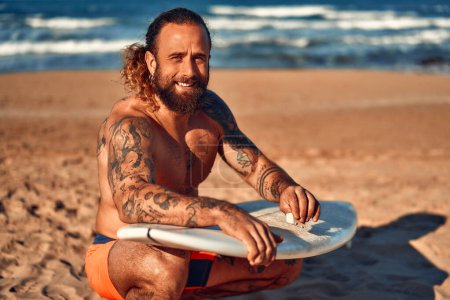 Téléchargez les photos : Homme barbu caucasien costaud avec des tatouages dans les maillots de bain cire une planche de surf près de la mer. Sports et loisirs actifs. - en image libre de droit