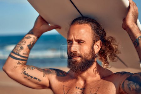 Foto de Caucásico surfista fornido barbudo hombre con tatuajes y pelo largo en baúles de natación con una tabla de surf cerca del mar. Deportes y recreación activa. - Imagen libre de derechos