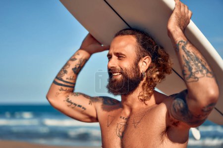 Photo for Caucasian beefy surfer bearded man with tattoos and long hair in swimming trunks with a surfboard near the sea. Sports and active recreation. - Royalty Free Image