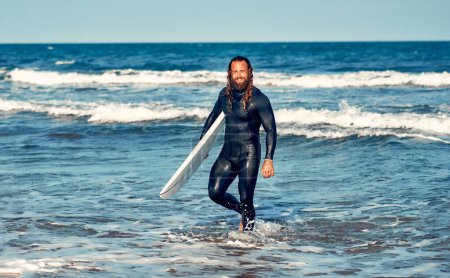Foto de Caucásico surfista fornido barbudo hombre con el pelo largo en un traje de neopreno con una tabla de surf cerca del mar. Deportes y recreación activa. - Imagen libre de derechos
