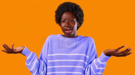 Foto de Mujer joven afroamericana con pelo afro peinado de pie en un suéter azul sobre un fondo naranja brillante extendió sus brazos a los lados en incredulidad. - Imagen libre de derechos