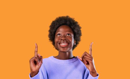 Téléchargez les photos : Afro-américaine jeune femme avec afro coiffure debout dans un pull bleu sur un fond orange vif pointe vers le haut avec un pouce vers le haut pour un produit publicitaire à insérer. - en image libre de droit