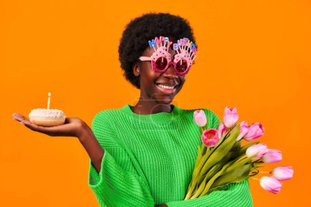 Photo for African american young woman with afro hair styling wearing funny glasses with happy birthday lettering standing in green sweater on bright orange background with cake with candle and tulips. - Royalty Free Image