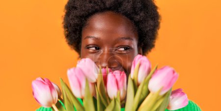 Foto de Mujer joven afroamericana con peinado afro de pie en un suéter azul sobre un fondo naranja brillante con un ramo de tulipanes. Día de la Mujer. - Imagen libre de derechos
