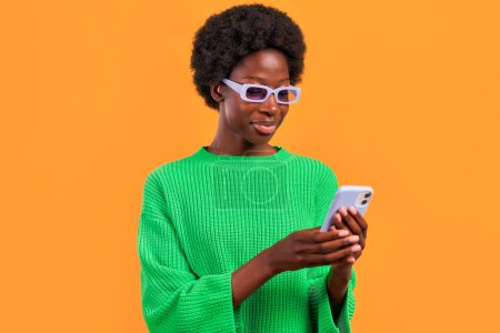 Téléchargez les photos : Afro-américaine jeune femme avec afro coiffure debout dans un pull vert sur un fond orange vif portant des lunettes et en utilisant le téléphone. - en image libre de droit