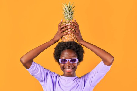 Téléchargez les photos : Afro-Américaine jeune femme avec coiffure afro portant des lunettes de soleil violettes tenant l'ananas isolé sur fond orange. - en image libre de droit