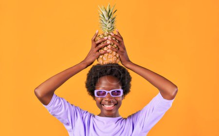 Photo for African American young woman with afro hair styling wearing purple sunglasses holding pineapple isolated on orange background. - Royalty Free Image