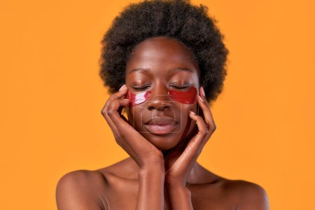 Foto de Mujer joven afroamericana con cabello afro haciendo su rutina matutina usando parches oculares hidratantes aislados sobre fondo naranja. Tratamientos de cuidado de la piel y spa. - Imagen libre de derechos
