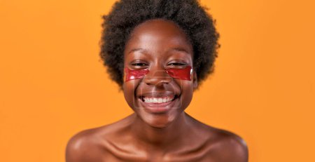 Téléchargez les photos : Jeune femme afro-américaine aux cheveux afro faisant sa routine matinale en utilisant des patchs hydratants pour les yeux isolés sur fond orange. Soins de la peau et soins spa. - en image libre de droit
