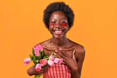Photo for African American young woman with afro hair using moisturizing eye patches holding bouquet of tulips isolated on orange background. Skin care and spa treatments. - Royalty Free Image