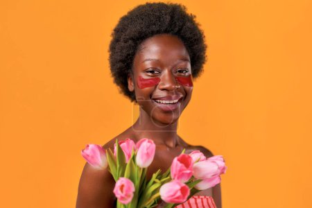 Photo for African American young woman with afro hair using moisturizing eye patches holding bouquet of tulips isolated on orange background. Skin care and spa treatments. - Royalty Free Image