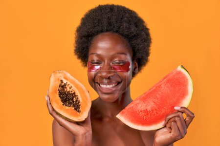 Téléchargez les photos : Jeune femme afro-américaine aux cheveux afro en utilisant des patchs hydratants pour les yeux, tenant papaye et pastèque dans les mains isolées sur fond orange. Soins de la peau et soins spa. - en image libre de droit
