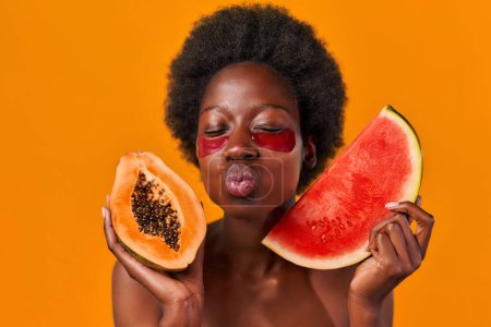 Téléchargez les photos : Jeune femme afro-américaine aux cheveux afro en utilisant des patchs hydratants pour les yeux, tenant papaye et pastèque dans les mains isolées sur fond orange. Soins de la peau et soins spa. - en image libre de droit