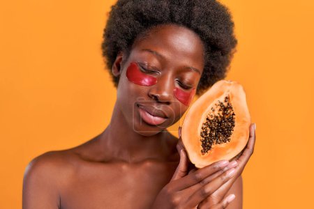 Téléchargez les photos : Jeune femme afro-américaine aux cheveux afro utilisant des patchs hydratants tenant la papaye dans ses mains isolées sur fond orange. Soins de la peau et soins spa. - en image libre de droit
