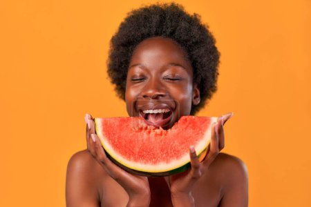 Photo for African American young woman with afro hair takes a tasty bite of juicy watermelon isolated on orange background. Skin care. Healthy lifestyle. - Royalty Free Image