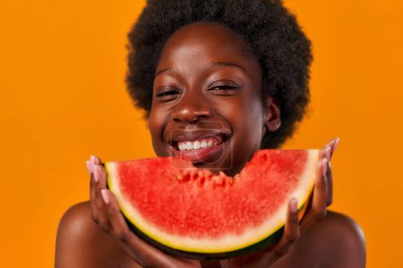 Photo for African American young woman with afro hair takes a tasty bite of juicy watermelon isolated on orange background. Skin care. Healthy lifestyle. - Royalty Free Image