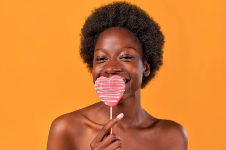Téléchargez les photos : Afro-américaine jeune femme aux cheveux afro tenant des cœurs de bonbons roses isolés sur fond orange. Saint Valentin. - en image libre de droit