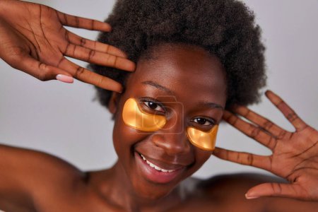 Foto de Mujer joven afroamericana con pelo afro y perfecta incluso la piel sana usando parches para los ojos aislados sobre fondo gris. Tratamientos de cuidado de la piel y spa. - Imagen libre de derechos