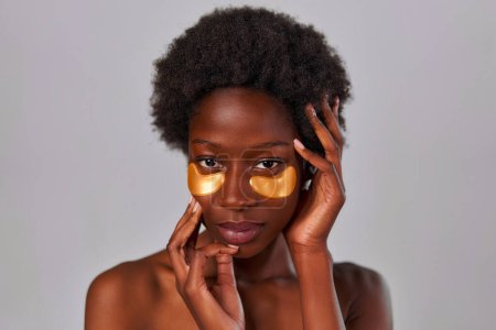Foto de Mujer joven afroamericana con pelo afro y perfecta incluso la piel sana usando parches para los ojos aislados sobre fondo gris. Tratamientos de cuidado de la piel y spa. - Imagen libre de derechos