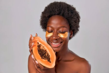 Téléchargez les photos : Afro-Américaine jeune femme avec des cheveux afro et une peau lisse et saine parfaite en utilisant des patchs pour les yeux et en tenant la papaye dans les mains isolées sur fond gris. Soins de la peau et soins spa. - en image libre de droit