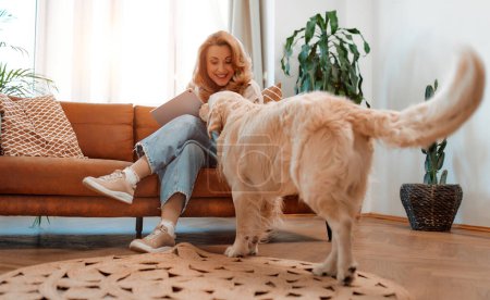 Téléchargez les photos : Jolie femme blonde assise sur un canapé avec une tablette dans le salon à la maison avec son chien labrador, s'amuser et passer du temps ensemble. - en image libre de droit