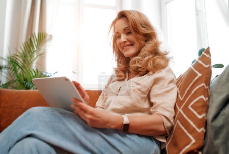 Téléchargez les photos : Belle femme blonde assise sur le canapé avec une tablette dans le salon à la maison, faisant des achats en ligne, travaillant ou bavardant en ligne. - en image libre de droit