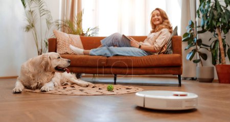 Téléchargez les photos : Une belle femme blonde est assise sur un canapé avec une tablette dans le salon à la maison avec un chien labrador, au premier plan un robot aspirateur nettoie la maison. - en image libre de droit