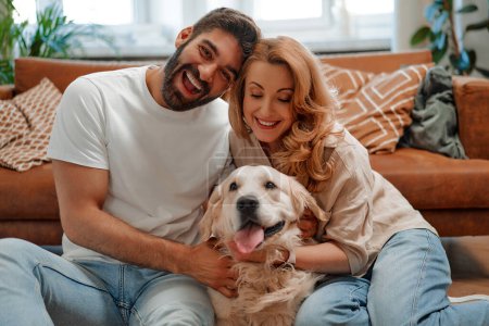 Téléchargez les photos : Jeune couple jouant et embrassant leur chien labrador animal de compagnie assis sur le canapé près de la fenêtre dans le salon à la maison, s'amusant ensemble. - en image libre de droit