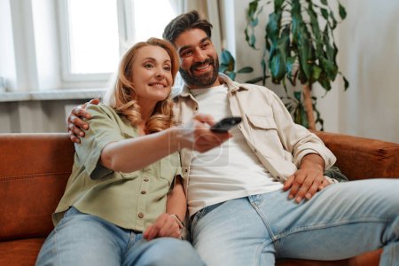 Téléchargez les photos : Couple adulte avec télécommande regarder un film à la télévision tout en s'amusant assis sur le canapé près de la fenêtre dans le salon à la maison. - en image libre de droit