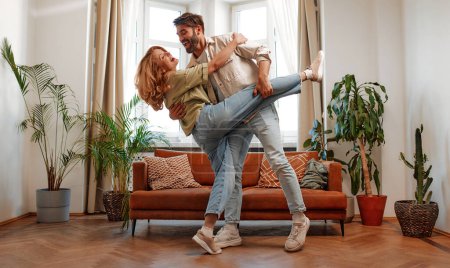Téléchargez les photos : Un couple d'adultes amoureux dansant joyeusement dans le salon sur le fond du canapé et de la fenêtre à la maison, s'amusant et romantique ensemble. - en image libre de droit